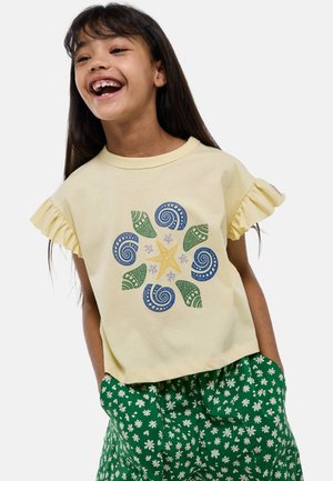 T-shirt jaune à manches courtes avec des bords froncés, ornée d'un motif de coquillage multicolore et d'étoiles de mer. Associée à un short vert à motif floral.