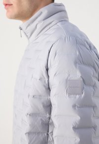 Lichtgrijze pufferjack met een staande kraag, gestikt patroon en een klein vierkant logo-opdruk op de linker mouw.