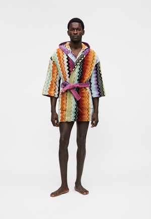 Homme debout pieds nus portant une robe courte à capuche avec des motifs zigzag colorés et une ceinture rose nouée sur un fond uni.