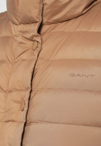 Bēšs polsterēts veste ar augstu apkakli, spīdīgu tekstūru un pogu aizdari. Uz auduma ir diskrēti uzdrukāts zīmola nosaukums "GANT".