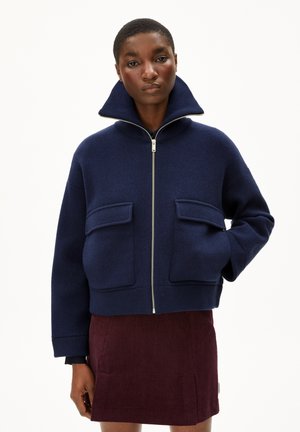 Marineblauwe wolmix ritsjack met hoge kraag, voorzien van twee voorzakken en een ontspannen pasvorm. Textuurstof en stevige ritsdetail.