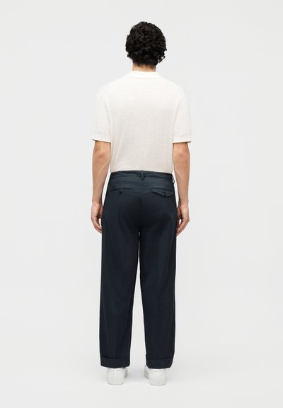 Homme aux cheveux bouclés portant une chemise blanche à manches courtes, un pantalon bleu marine avec poches arrière et des baskets blanches, debout de dos.