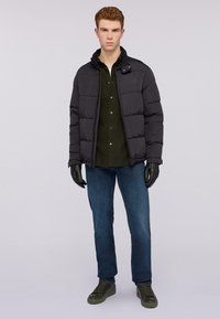 Chaqueta puffy negra con cuello alto, llevada sobre una camisa verde oscura, combinada con vaqueros azules y guantes negros, con zapatillas verdes.