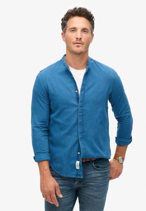Chemise en denim bleu profond, dotée d'un col mandarin, d'une fermeture à boutons, de manches retroussées et d'un ourlet droit. Portée par-dessus un t-shirt blanc.