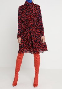 Red and black floral patterned dress with long sleeves, gathered waist, and knee-length skirt, paired with glossy red over-the-knee boots.