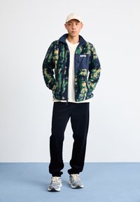 Fleecejacka med gröna, gula och marinmönster, full dragkedja, ståkrage och bröstficka. Sammanställd med svarta byxor och sneakers.
