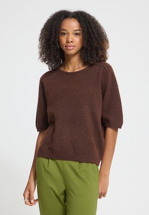 Jeune femme aux cheveux bouclés portant un pull marron à manches courtes et un pantalon vert, debout devant un fond blanc uni.