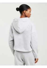 Žena s tamnom kosom u punđi nosi svijetlosivu hoodicu i odgovarajuće trenirke, stojeći okrenuta leđima prema jednostavnoj svijetloj pozadini.