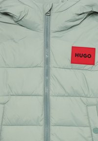 Šviesiai žalia pūkinė striukė, su priekiniais užtrauktukais, horizontaliu siuvinėjimu ir raudonu stačiakampiu žyma "HUGO" juodu.