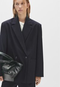 Blazer bleumarin cu două rânduri de nasturi, dungi subțiri, revere decupate, doi nasturi și buzunare laterale, asociat cu o geantă neagră texturată.