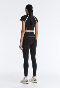Femme debout de dos portant un crop top noir ajusté et des leggings avec des détails de couture blancs, ainsi que des baskets blanches sur un fond uni.