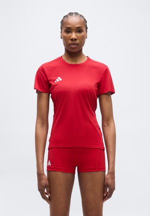 Femme vêtue d'un t-shirt de sport rouge et d'un short, debout droite sur un fond blanc uni.
