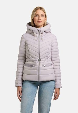 Junge Frau trägt eine hellviolette gesteppte Kapuzenjacke mit Gürtel und Reißverschlusstaschen, kombiniert mit hellblauen Jeans, steht vor weißem Hintergrund.