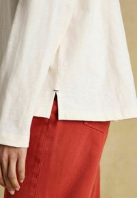 Külgvaade inimesest, kes kannab valget pikka varrukatoppi väikese küljeaugu ja punaste pükste ning sõrmi lõdvalt külje ääres.