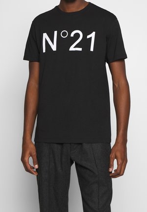 Personne portant un t-shirt noir à manches courtes avec un texte blanc "N°21" sur le devant et un pantalon gris foncé sur un fond clair uni.