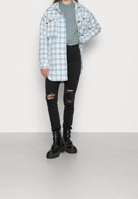 Donna vestita con un giubbotto a quadri blu, un maglione grigio a coste, jeans skinny strappati neri e stivaletti militari neri con fibbie, in piedi contro un muro bianco.