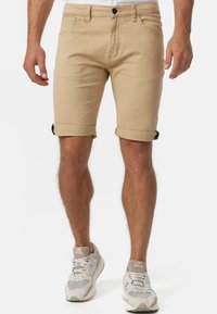 Beige bomullsshorts med valsade fållar, med framfickor och knäppning. Bärs med grå och vita sneakers.