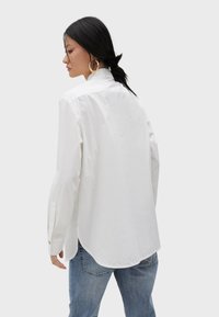 White cotton long-sleeve shirt with a buttoned collar, a slightly curved hem, and cuffs. Paired with light blue denim jeans.