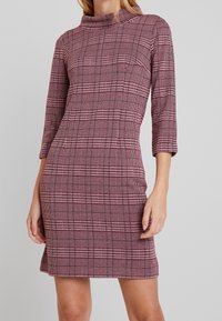 Punainen ruudullinen mekko, jossa on tiukka siluetti, korkeampi kaula-aukko ja kolme neljäsosaa hihat. Teksturoitu kangas esittelee ruutukuosin.