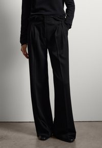 Zwarte wijde broek met plooien aan de voorkant en een gladde textuur, gecombineerd met een aansluitende zwarte trui. De schoenen hebben een slank ontwerp.
