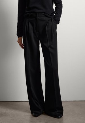 Zwarte wijde broek met plooien aan de voorkant en een gladde textuur, gecombineerd met een aansluitende zwarte trui. De schoenen hebben een slank ontwerp.