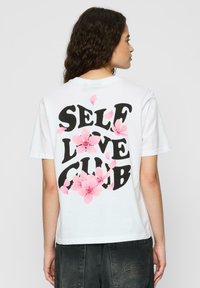 Tricou alb din bumbac cu "SELF LOVE CLUB" imprimat cu litere negre îndrăznețe pe spate, decorat cu desene florale roz și petale.