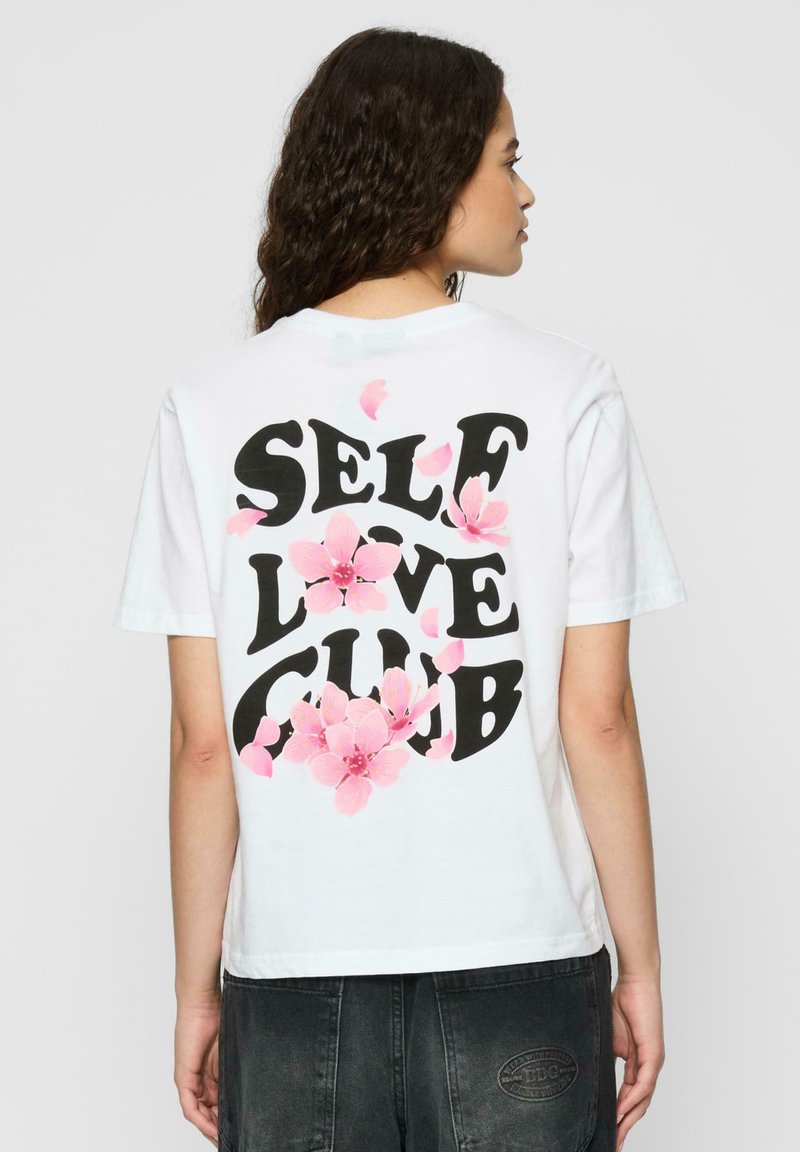 Tricou alb din bumbac cu "SELF LOVE CLUB" imprimat cu litere negre îndrăznețe pe spate, decorat cu desene florale roz și petale.