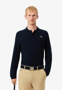 Navy lange mouw polo met een knoopsluiting en groen geborduurd logo; gecombineerd met beige broek en een bijpassende riem.