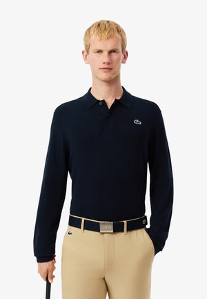 Navyblaues Langarm-Polohemd mit einem knopfbaren Kragen und grünem gesticktem Logo; kombiniert mit beigen Hosen und einem passenden Gürtel.