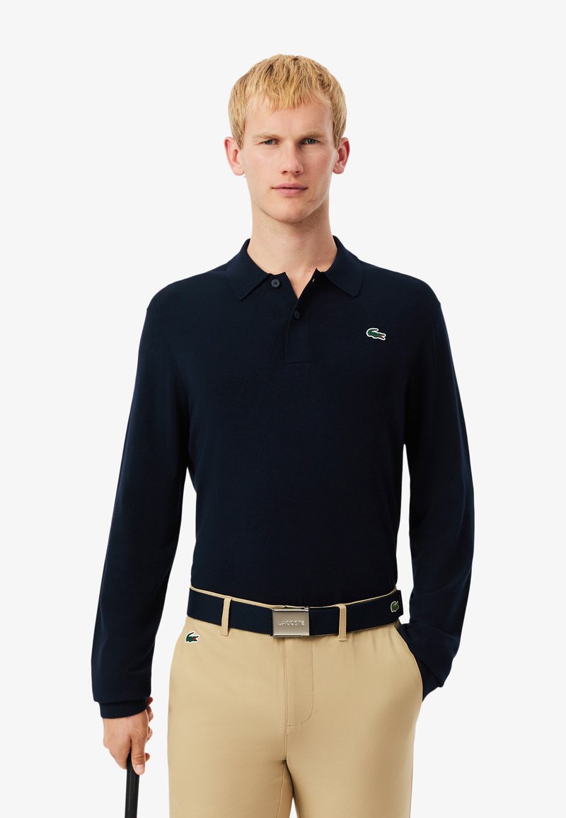 Navy lange mouw polo met een knoopsluiting en groen geborduurd logo; gecombineerd met beige broek en een bijpassende riem.