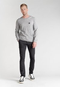Camicia grigia a maniche lunghe con un piccolo logo nero, abbinata a pantaloni neri slim-fit e sneakers nere/bianco, in piedi davanti a uno sfondo chiaro.