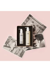 Twee flessen in een geschenkdoos: één grijs, één beige, beide met pompdispenser. De doos heeft een zwarte inzet. Op de achtergrond zijn vintage foto’s te zien.