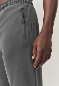 Gray athletic pants with an elastic waistband, side zip pocket, and textured logo patch on thigh. Smooth fabric, casual fit.