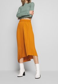 Jupe midi plissée orange avec une taille élastique, associée à un haut à manches longues vert et des bottines blanches à talon carré.