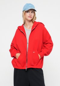 Felpa rossa con cappuccio zipato, dotata di un grande cappuccio e tasche frontali, realizzata in un materiale morbido. Presenta un piccolo logo bianco sul lato sinistro.