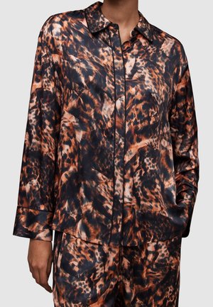 Personne portant une chemise à manches longues et un pantalon avec un motif abstrait d'animal orange, noir et marron, main dans la poche.