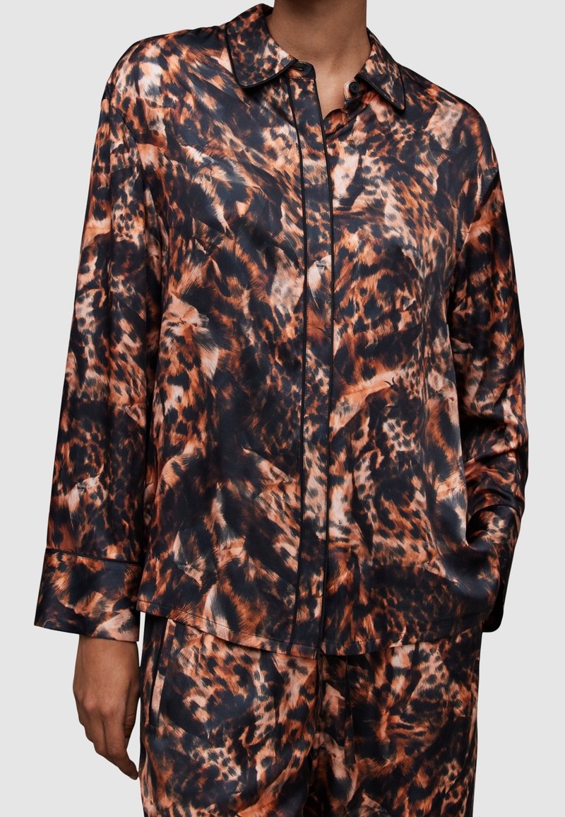 Personne portant une chemise à manches longues et un pantalon avec un motif abstrait d'animal orange, noir et marron, main dans la poche.