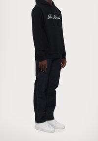 Muž v černé mikině s bílým textem, tmavě modrých džínách a bílých teniskách, stojící na světlém jednobarevném pozadí.