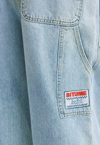 Veste en jean bleu clair avec une poche poitrine visible, des coutures dorées et un patch logo rouge et blanc portant l'inscription "BITUME solid".