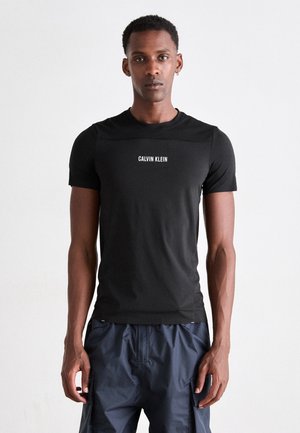 Man wearing black Calvin Klein short-sleeve shirt and dark blue cargo pants, standing against a plain light background.