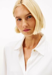 En blond kvinde med kort hår iført en hvid skjorte med krave, ser direkte frem med et neutralt udtryk foran en hvid baggrund.
