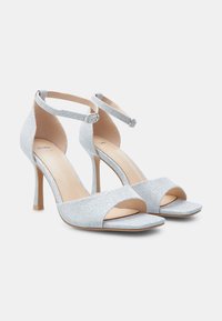 Sandalias de tacón alto plateadas brillantes con diseño de punta abierta, correa en el tobillo con hebilla y una forma elegante de tejido texturizado.