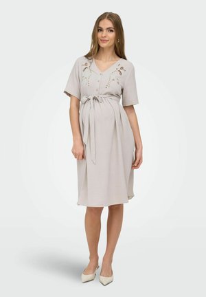 Jeune femme enceinte debout, portant une robe de maternité beige clair mi-genoux à manches courtes, boutonnée devant, avec des détails floraux découpés et une taille nouée.
