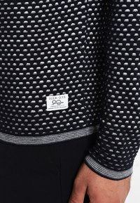 Chemise à manches longues à motifs noir et blanc avec un tissu texturé. Présente une étiquette portant la mention "PIER-ONE" et des poignets rayés.