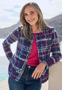 Gewebte Jacke mit einem Muster in Blau, Rot und Grau, runder Ausschnitt und Knopfverschluss, kombiniert mit einem rosafarbenen Oberteil und dunklen Jeans.