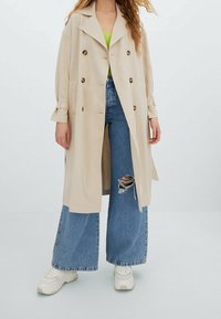 Manteau trench beige avec de grands boutons, poches à rabat et coupe décontractée. Porté sur un top vert et associé à un jean large et des baskets blanches.