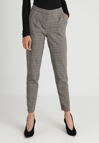 Szare spodnie w kratę z wysokim stanem, zaszewkami i zwężającymi się nogawkami, wykonane z gładkiego materiału. Połączone są z czarnymi szpilkami.