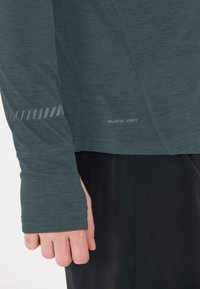 Langarm, donkergroene shirt van lichtgewicht stof met een gladde textuur. Kenmerkt zich door een reflecterend patroon op de mouw en een "QUICK DRY" label.