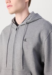 Grijze zip-up hoodie van zacht materiaal, met een trekkoord, een geborduurd logo op de borst en een relaxte pasvorm.