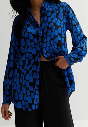 Vrouw draagt een blauw-zwart patroon blouse met lange mouwen en zwarte broek, trekt de blouse lichtjes omhoog aan één kant met haar hand.